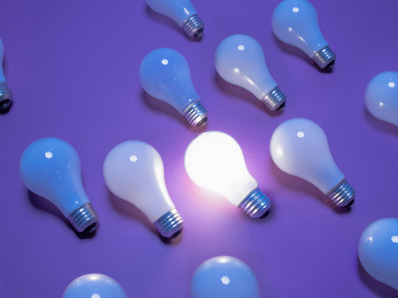 A group of lightbulbs lay on a purple background with only one lit up. 