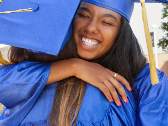 Two young women hug at high school graduation while wearing their caps and gowns. 