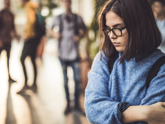 An uncertain young teen stands in the school hall with her arms wrapped around her. 