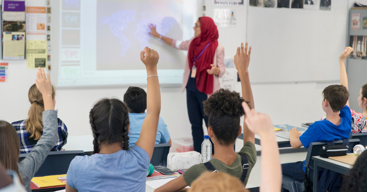 A stands teacher at the front of a classroom while students raise their hands. 