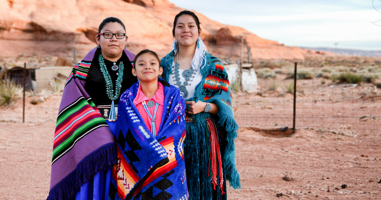 Three young Native women smile while wearing traditional clothes. 