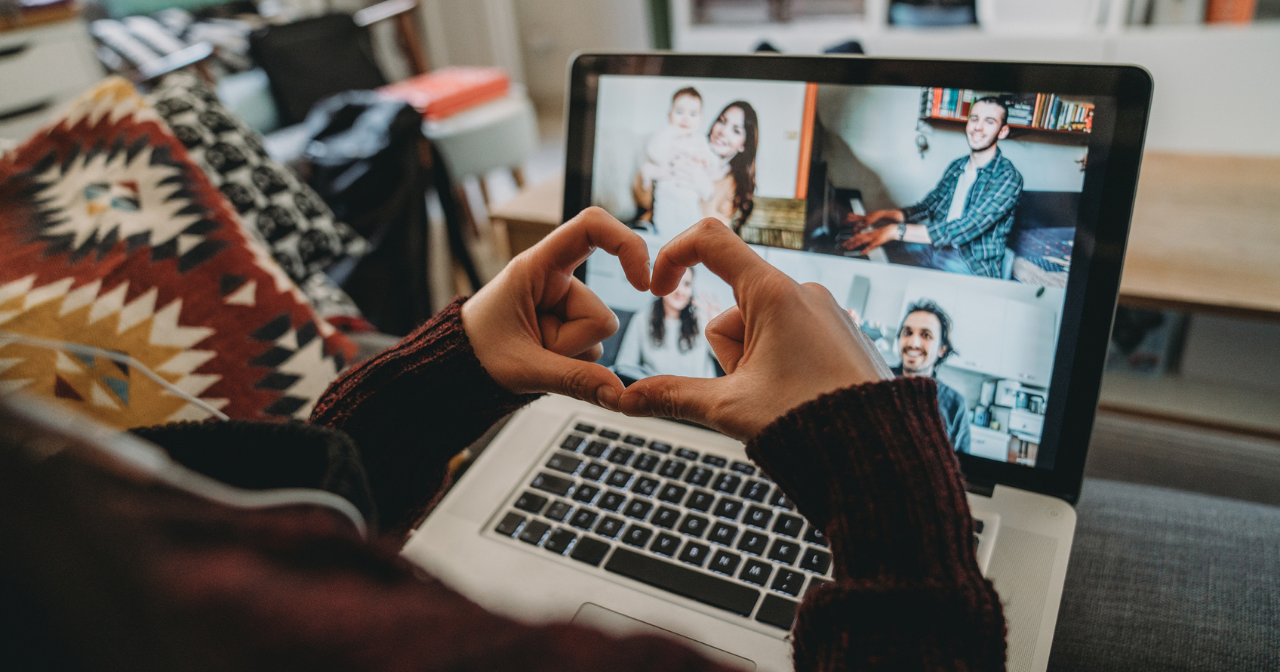 A person makes a heart with their fingers in front of their computer during a Zoom call with friends. 