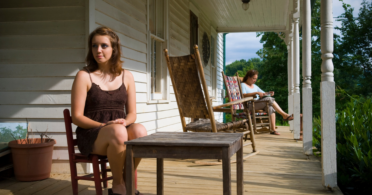 A young girl in Kentucky sits on her front porch with her mom in the background.