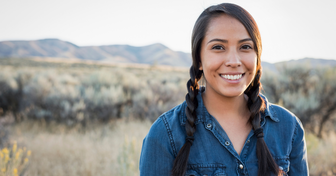 A young Native woman smiles for the camera while standing among a field of wild grasses. 