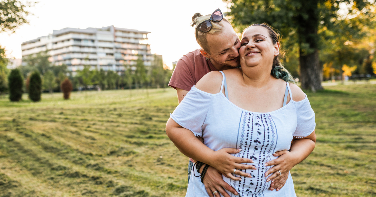 A pregnant woman smiles and is happily hugged by her male partner. 