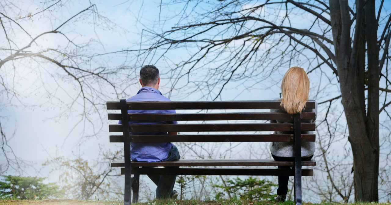 A couple sits on either end of a bench in a park. 