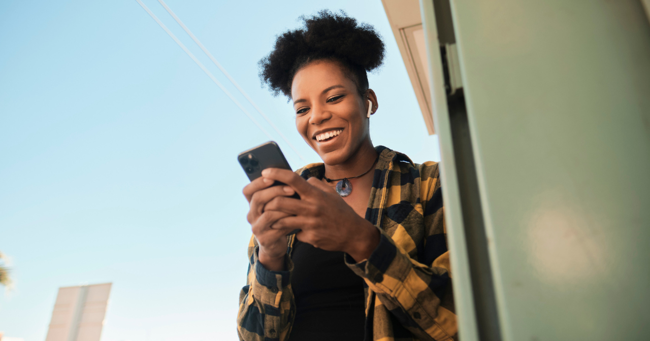 A woman leans against a wall outside and laughs while looking down at her phone. 