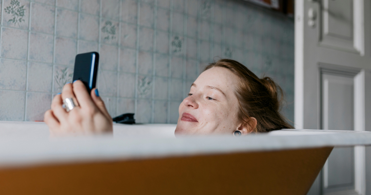A woman relaxes in a bath with a smile as she looks at her phone. 