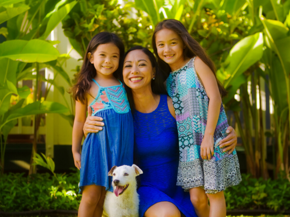 A mother, her two daughters, and a dog smile in a tropical setting.