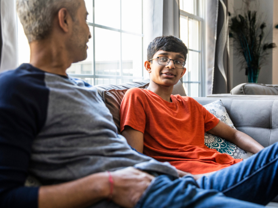 A father and his young teen son sit on the sofa and talk. 