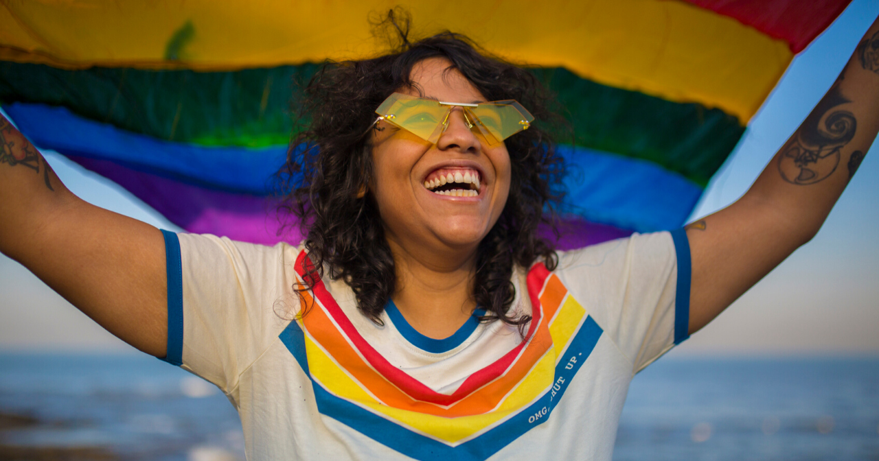 A woman in a rainbow tee holds a pride flag above her head and smiles