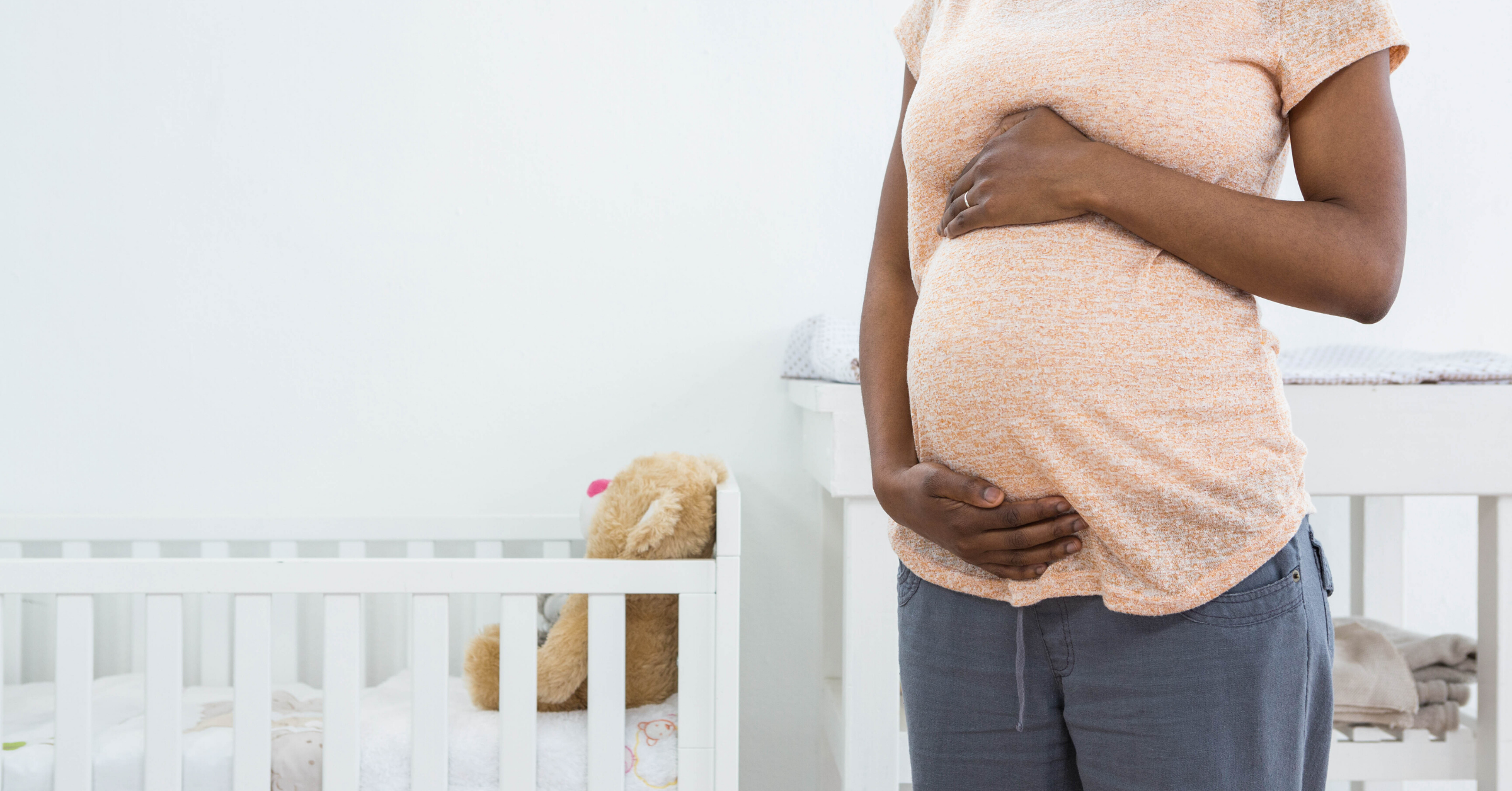 A pregnant Black woman holding her belly while standing next to a crib. 