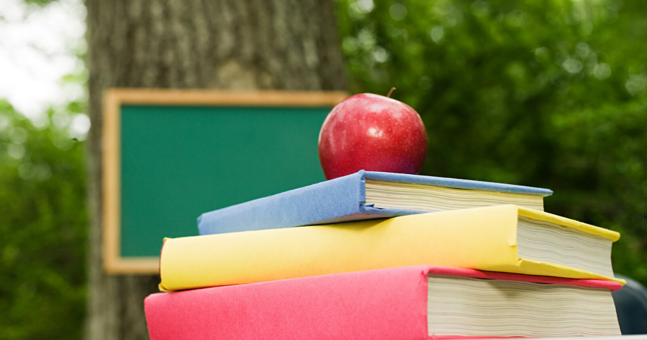 Outside, an apple sits on top of a pile of schoolbooks with a blackboard hung on a tree in the background. 