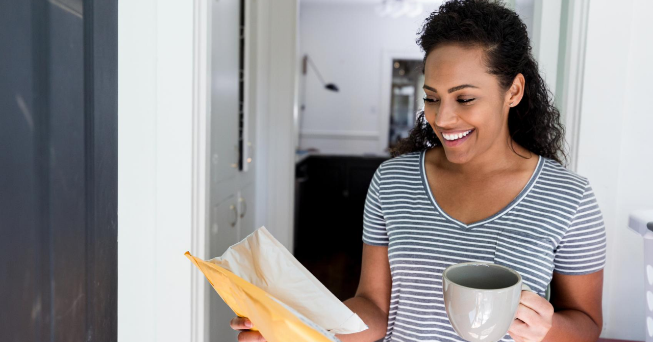 A woman holds a cup of coffee and with a smile picks up a package from her front door. 