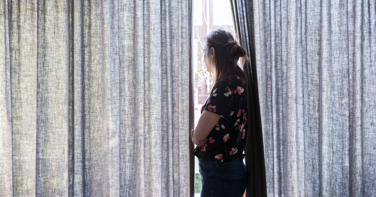 An image of a woman hugging herself and looking out of a window mostly covered by curtains. 