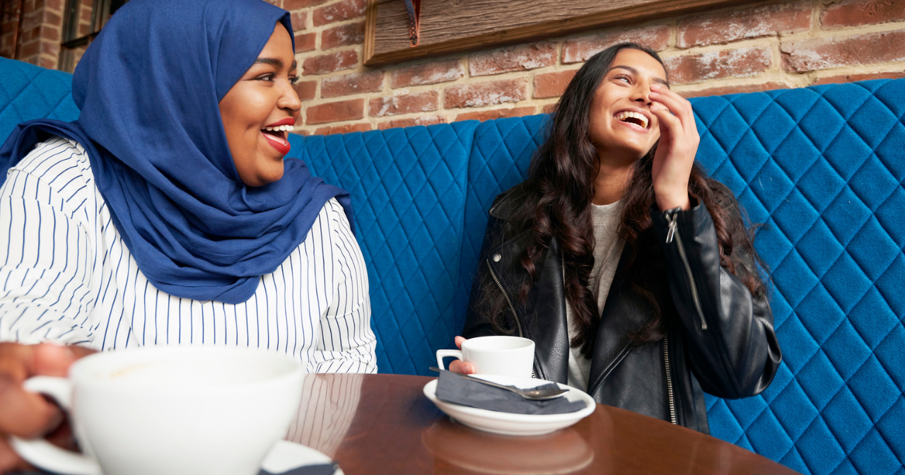 Two women sit at a table in a cafe and laugh over cups of coffee. 