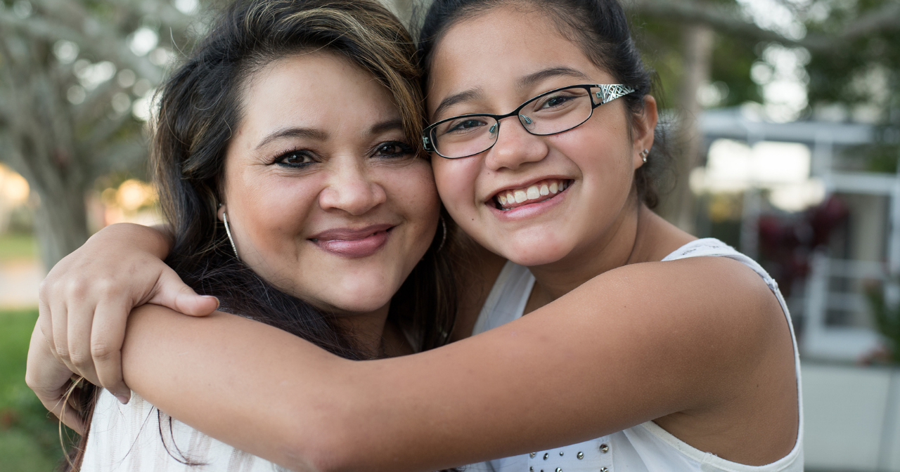 A Latina mom and her daughter hug and smile at the camera. 