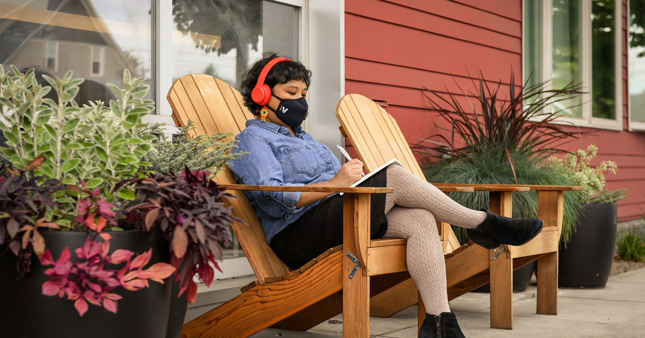 A Latinx person sits in a chair outside while wearing a facemask and writing a letter.
