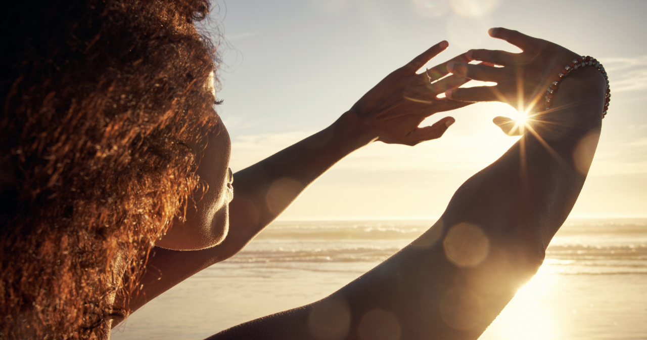 A Black woman stands on the beach and holds her hands up so she can look at the sunset. 
