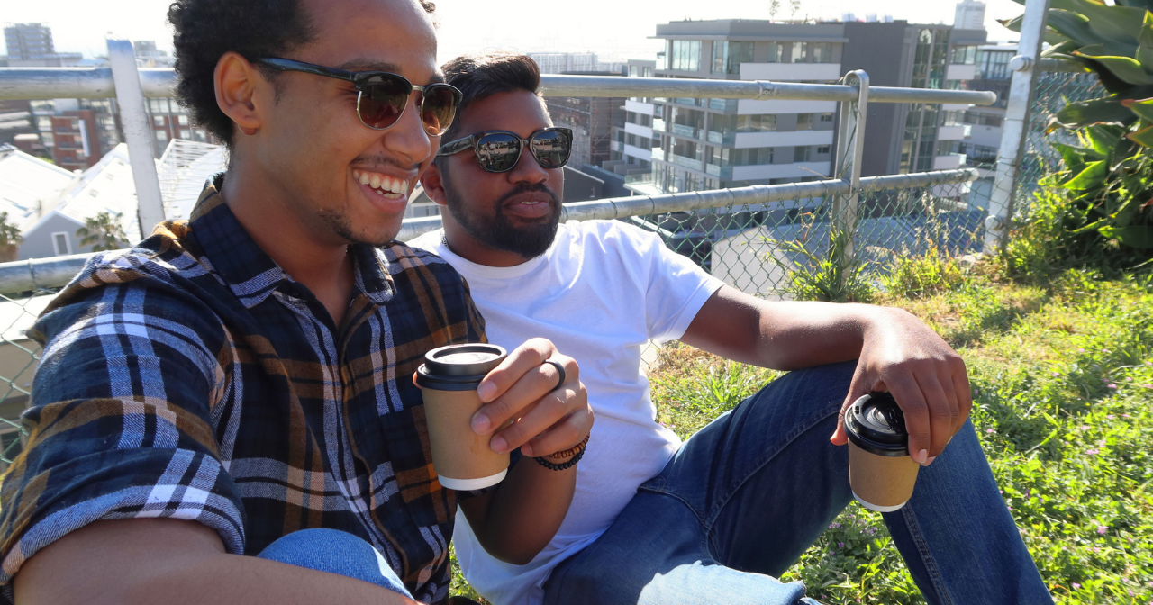 Two Black men relax on a rooftop with coffee. 