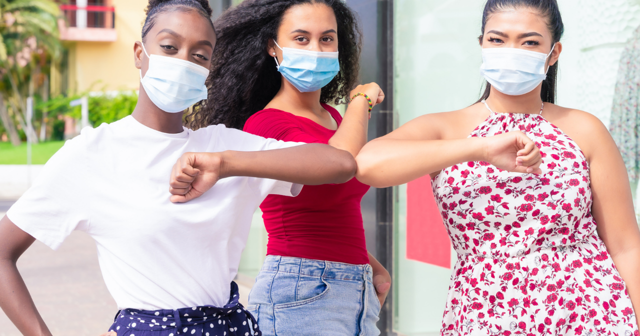 Three young women of color wear masks and touch elbows. 