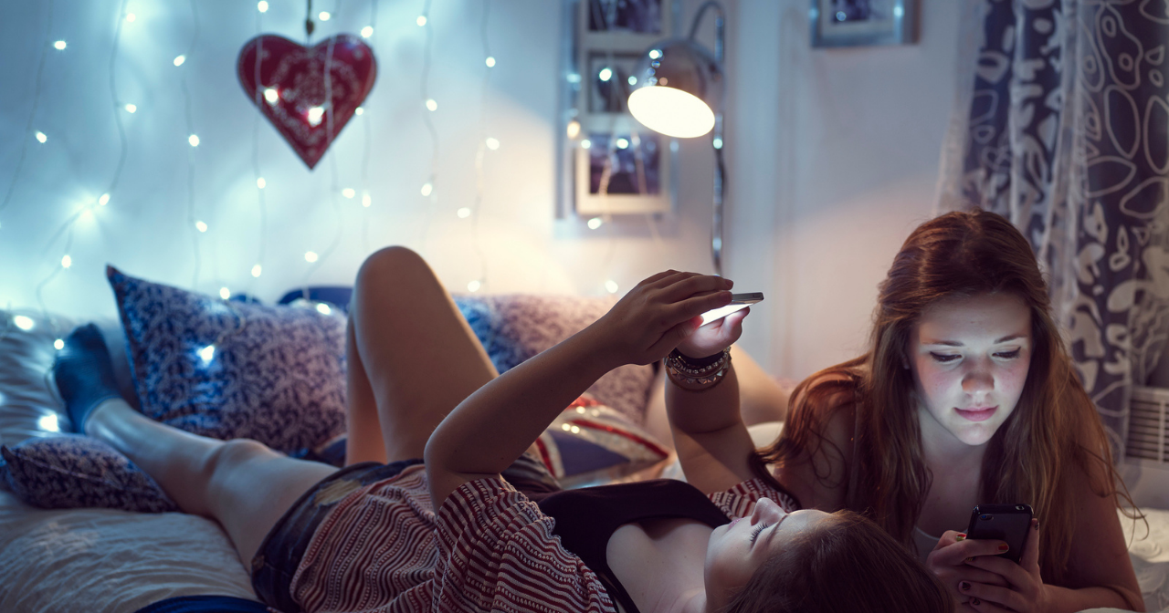 Two teen girls lay on a bed on their phones in soft lighting. 