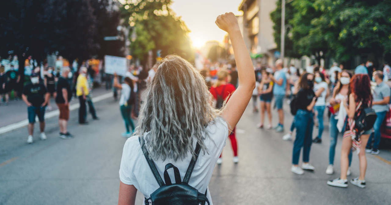 A photo of the back of a young woman at a protest with her fist raised. 
