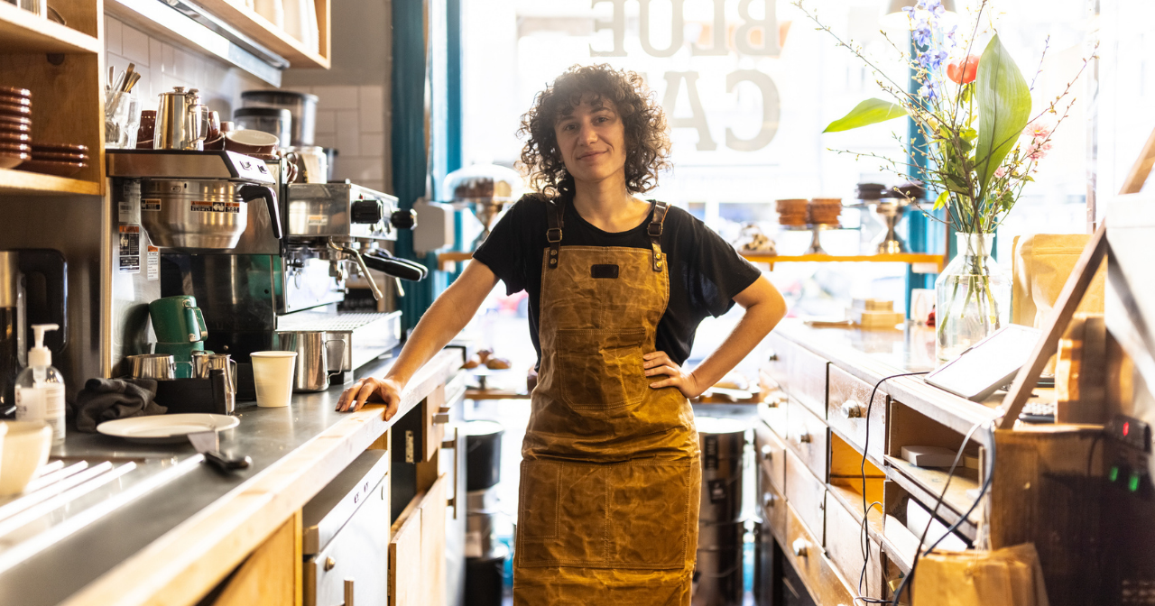 A person stands confidently behind the counter of the coffee that they work in. 