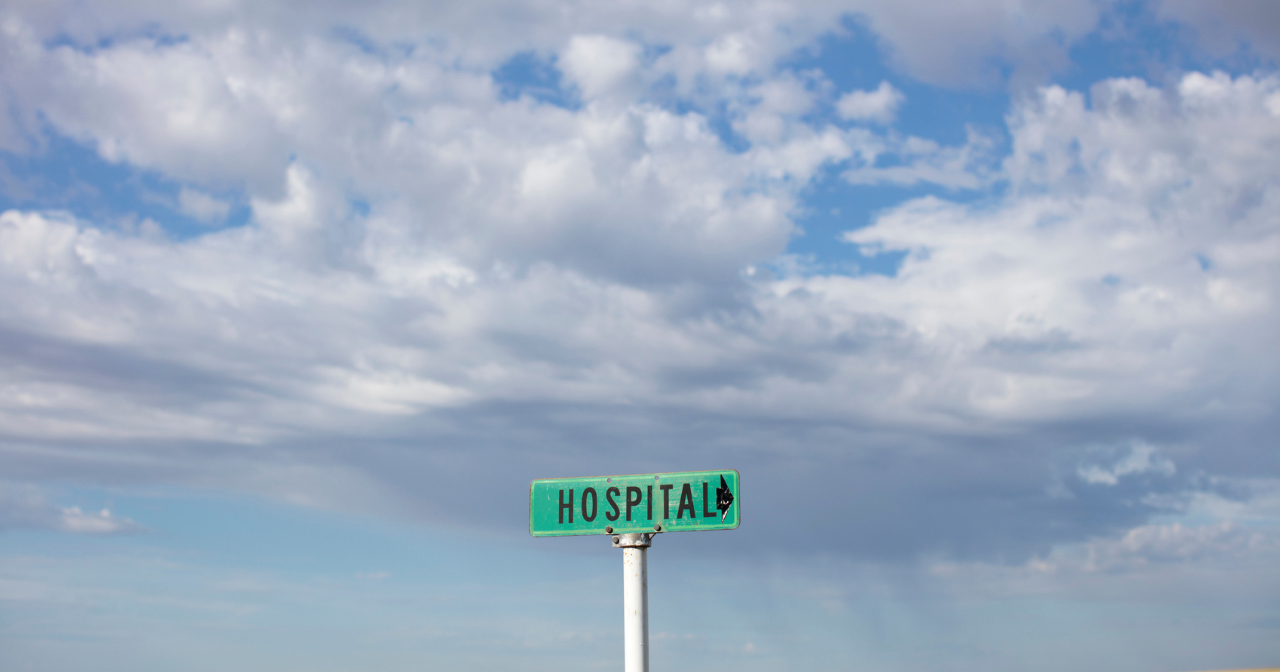 A green sign with the word hospital sits in the middle of a field against a blue sky. 