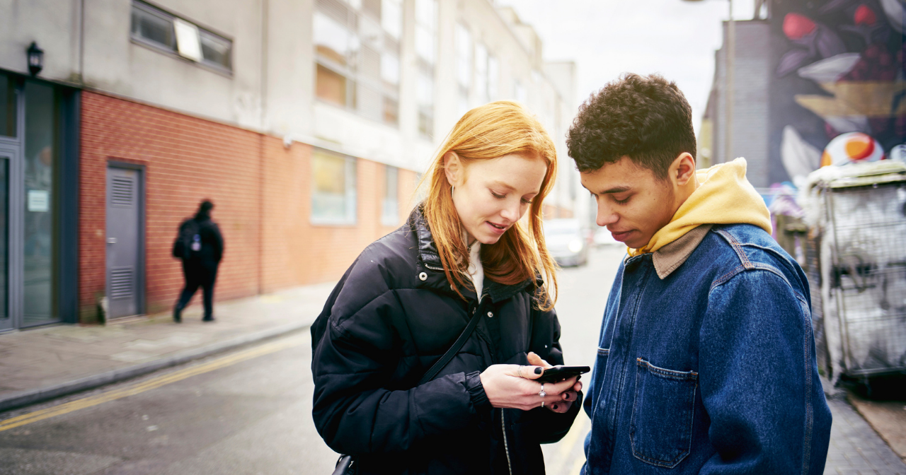 A young teen couple stands in the street and looks at a cell phone. 