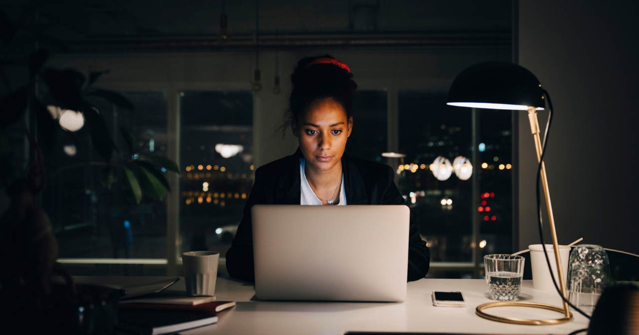 A woman sits at a desk on her computer late at night with only one light to work by. 