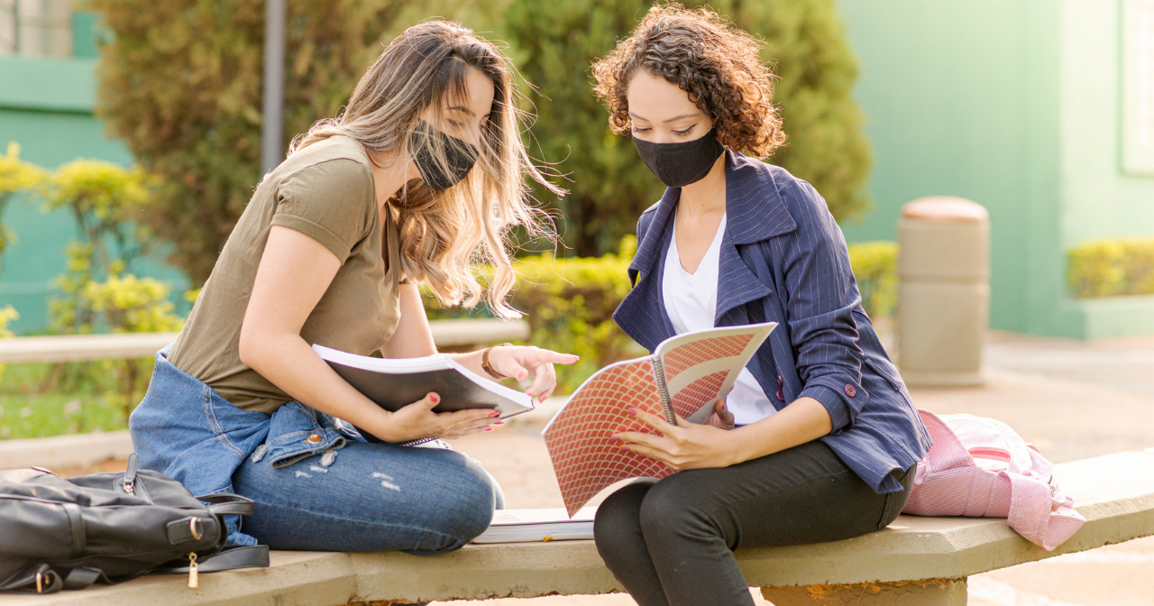 Two women wearing masks sit on a bench outside and study together. 