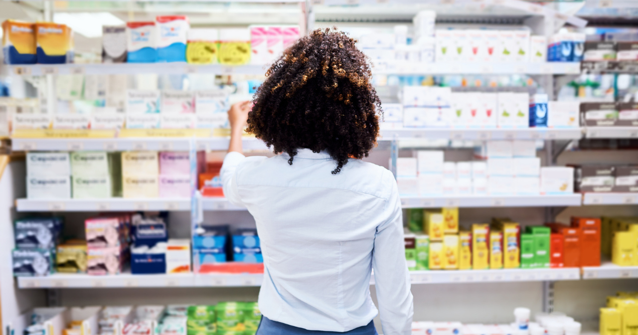 A young person stands with their back to the photo and considers a row of period products in a pharmacy. 