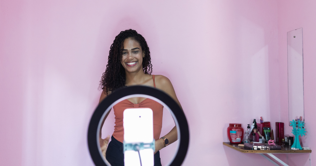 A woman poses in front of a pink wall with a phone set into a ring light in front of her, filming. 