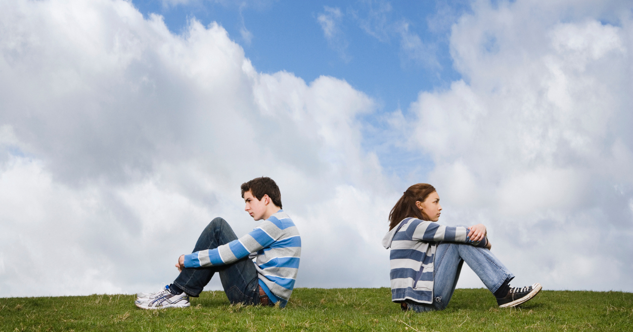 A teen couple sit on the top of a hill hugging their knees with their backs to one another. 