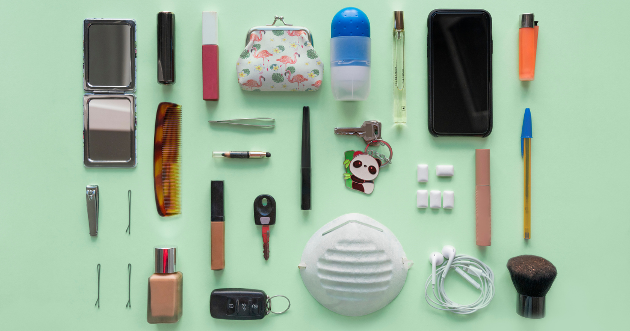 A green table with the contents of someone's bag laid out. Items include a mask, phone, comb, bobby pins, headphones, and more. 