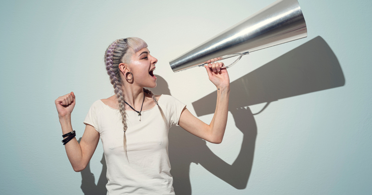 A woman holds a silver megaphone up to yell into against a blue background. 