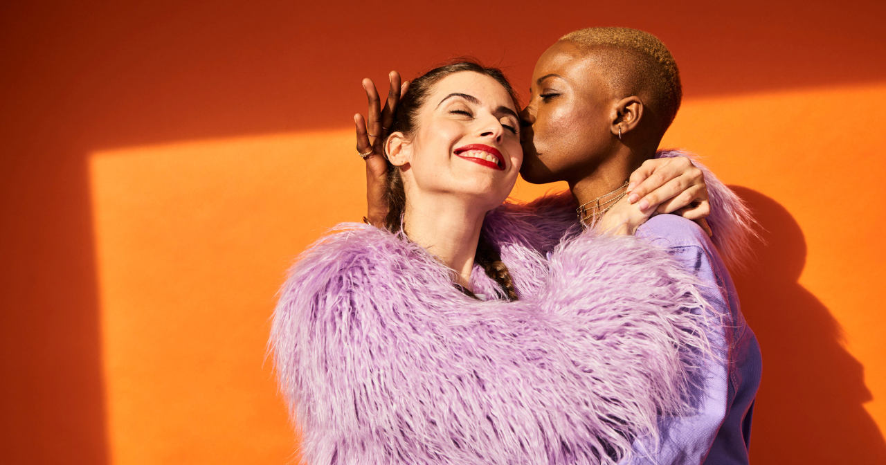 A woman kisses another woman's face while they hug against a bright orange background.