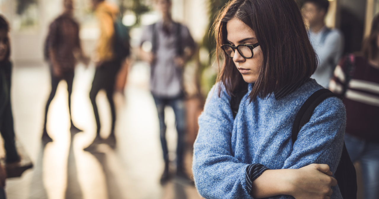 An uncertain young teen stands in the school hall with her arms wrapped around her. 