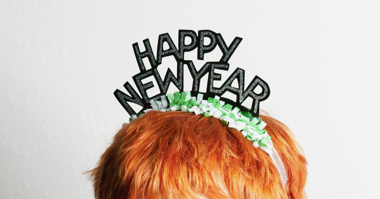 A photo of a the top of a person's head. They have red hair and are wearing a novelty headband that reads happy new year.