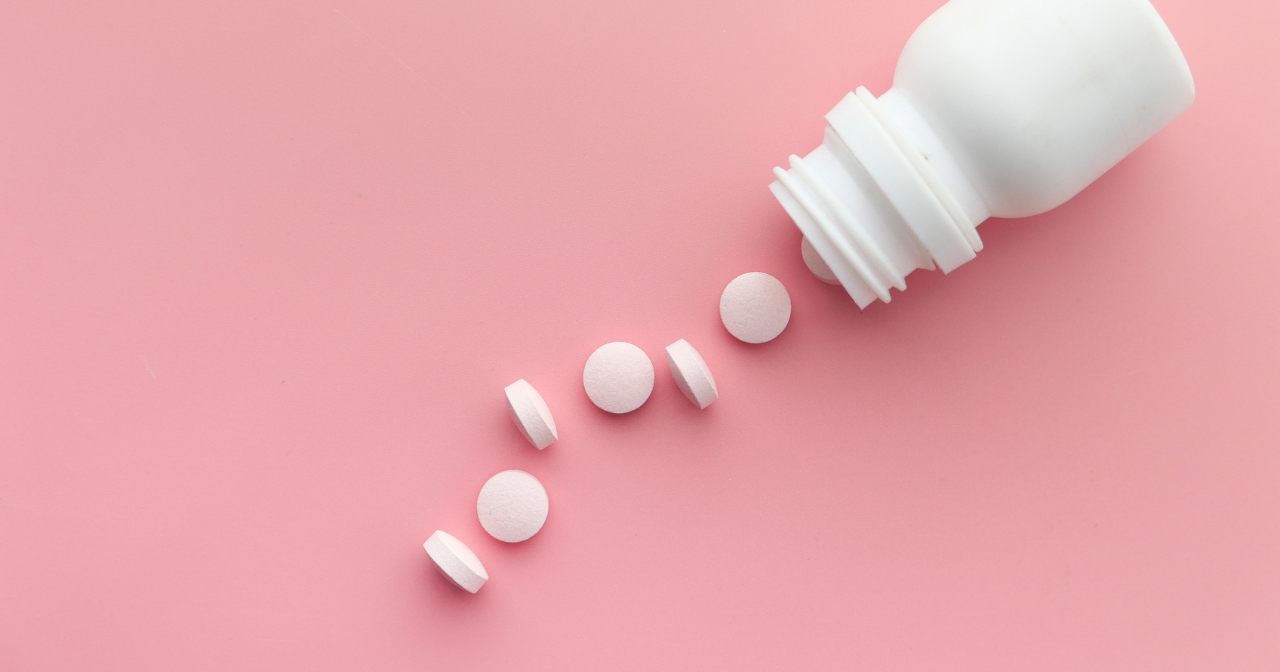A blank white bottle with small white pills spilling out on a pink background. 