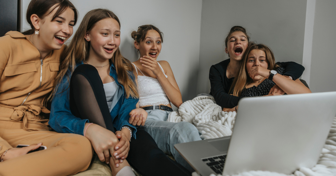 A group of 5 teen girls watch TV together and laugh wildly. 