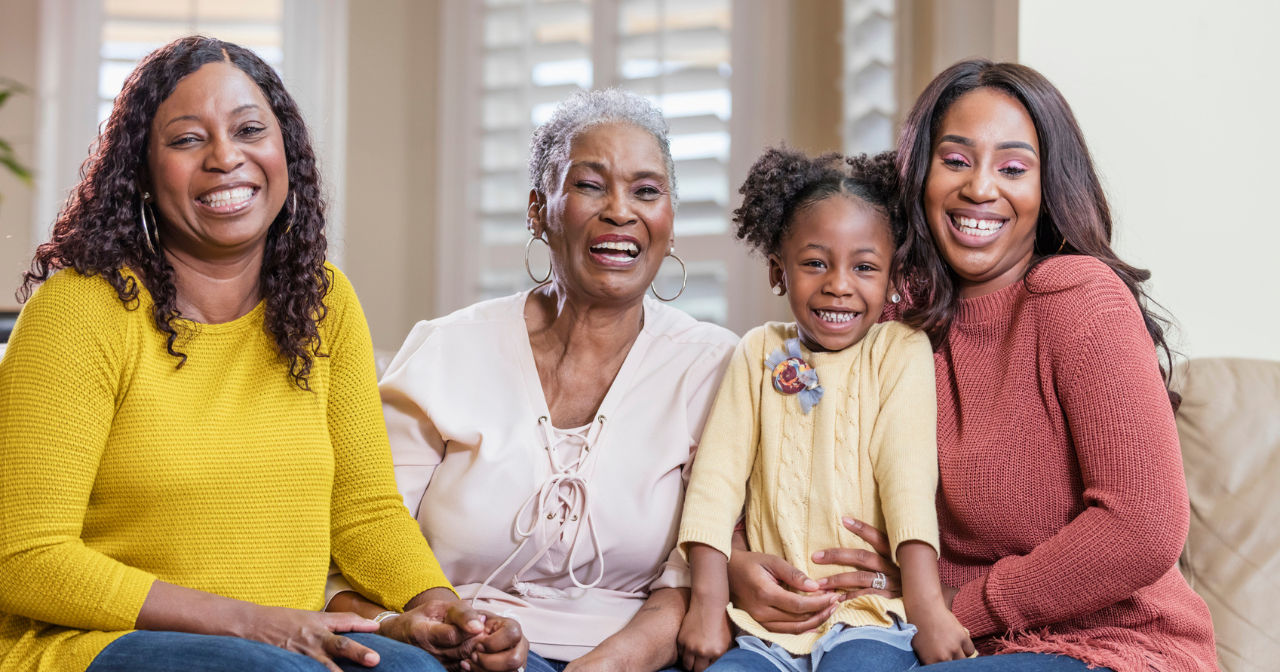 Four generations of Black women sit on a sofa and smile at the camera.