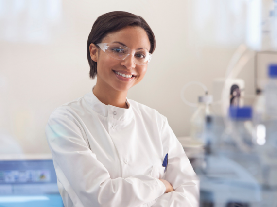 A woman in a white lab coat stands and smiles in her lab