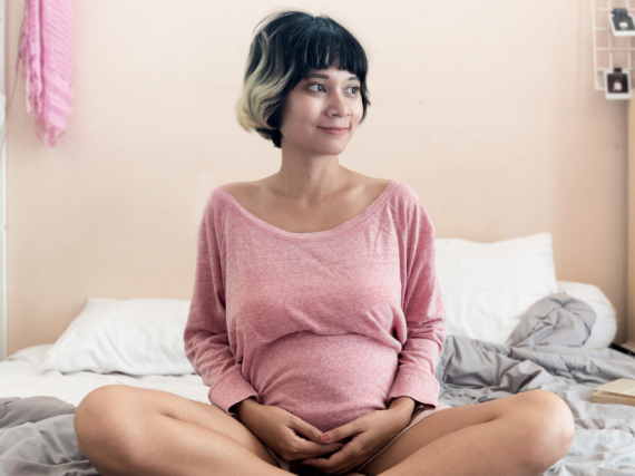 A pregnant woman sits cross legged on a bed