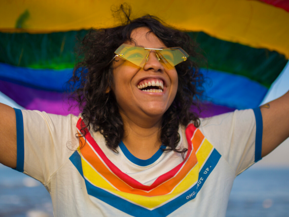 A woman in a rainbow tee holds a pride flag above her head and smiles