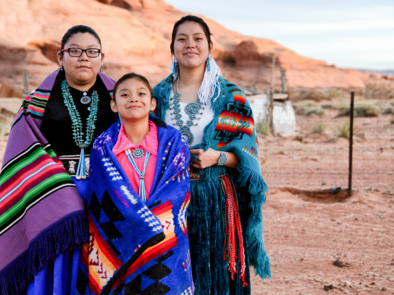 Three young Native women smile while wearing traditional clothes. 