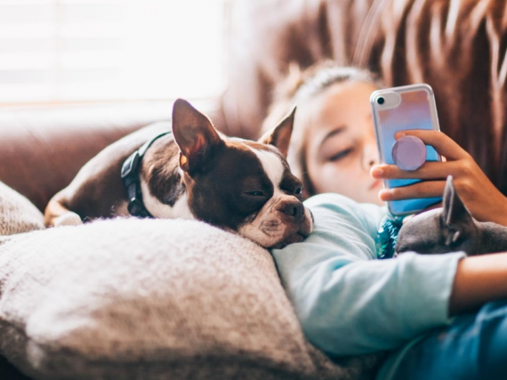 A young teen lays on the sofa with her dog while scrolling on her phone. 