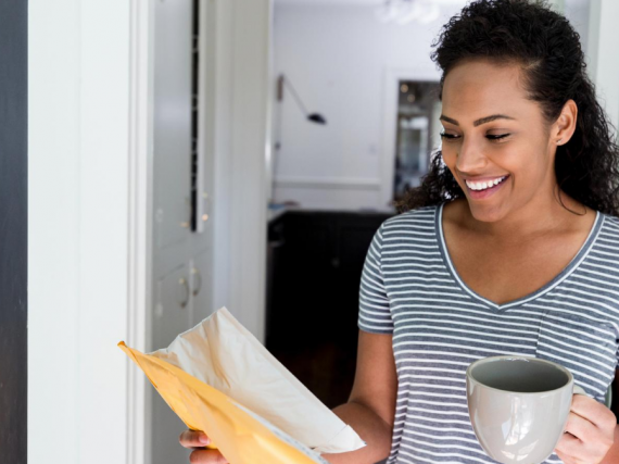 A woman holds a cup of coffee and with a smile picks up a package from her front door. 