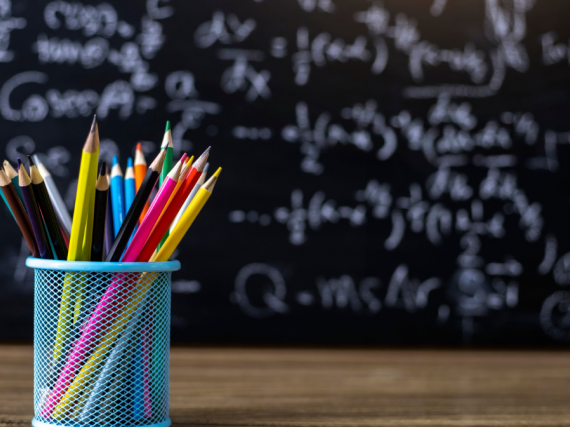 A container of colored pencils sit on a desk in front of a blackboard covered in chalk writing. 